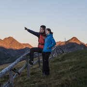 Zwei Wanderer stehen auf einer grünen Anhöhe und zeigen zu den von der Sonne beleuchteten Gipfeln.