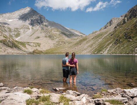 Paar steht in einem klaren Bergsee vor felsigen Alpengipfeln und blauem Himmel.
