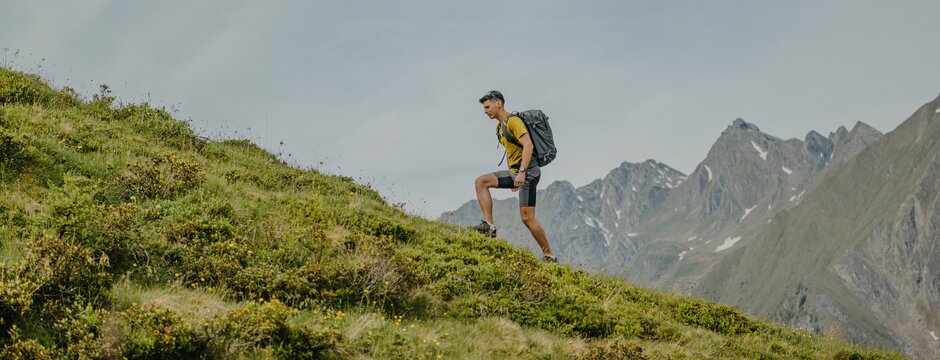 Escursionista che sale un pendio erboso alpino con le montagne sullo sfondo.