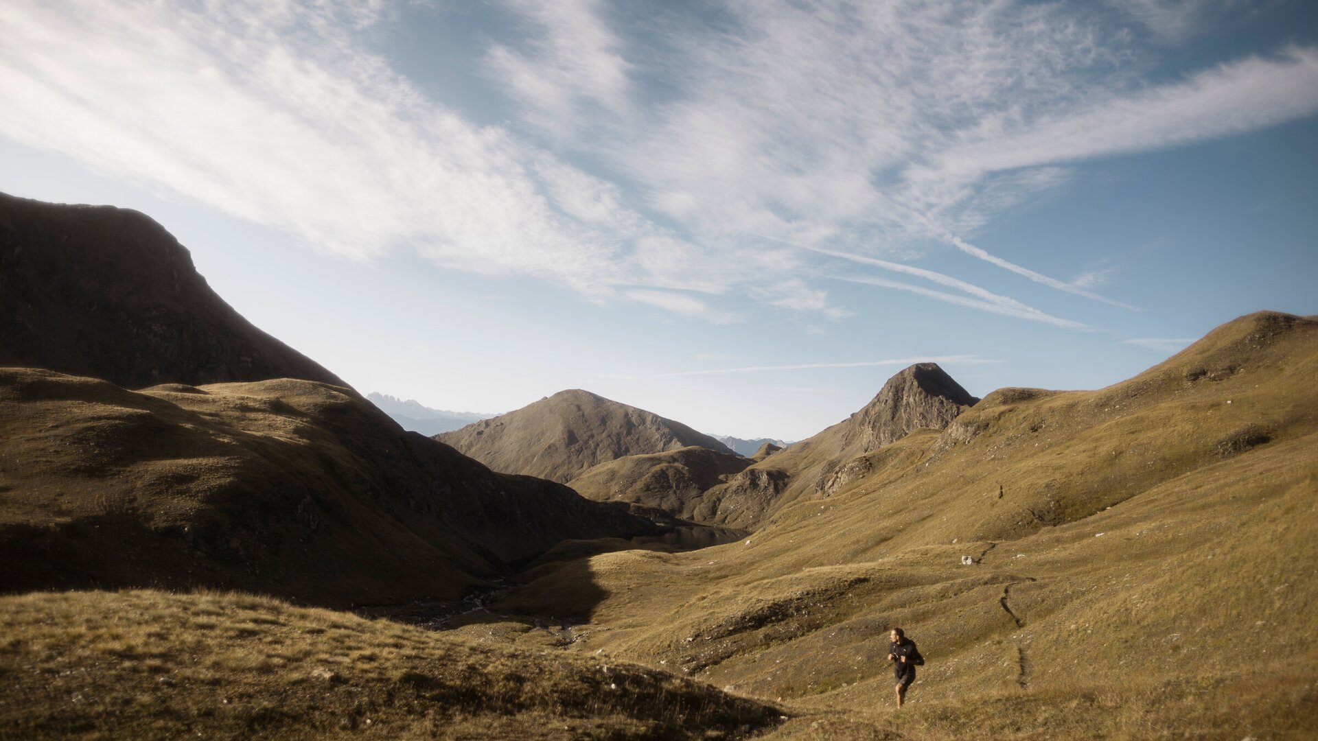 Hiker on a grassy mountain trail in a wide alpine valley under a blue sky. | © IDM Südtirol-Alto Adige/Manuel Kottersteger