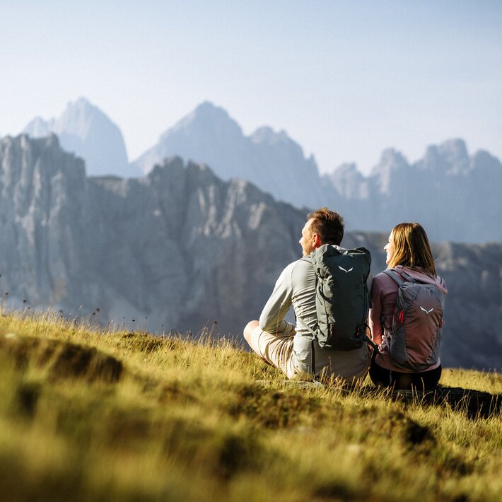 Zwei Wanderer sitzen auf einer Wiese mit markanten Gipfeln im Hintergrund.