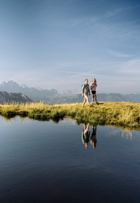 Zwei Wanderer gehen auf einem grasigen Grat am Bergsee mit spiegelnden Gipfeln entlang.
