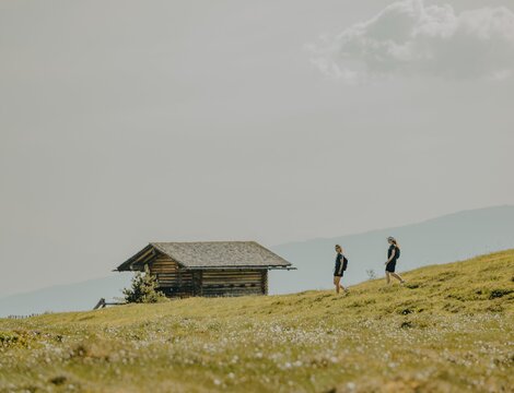 Zwei Wanderer an einer kleinen Almhütte auf einer grünen Bergwiese unter bewölktem Himmel.