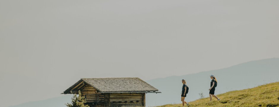 Due escursionisti vicino a una piccola baita alpina su un prato di montagna sotto un cielo nuvoloso.