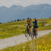 Two mountain bikers ride along a gravel path through an alpine meadow with wildflowers.