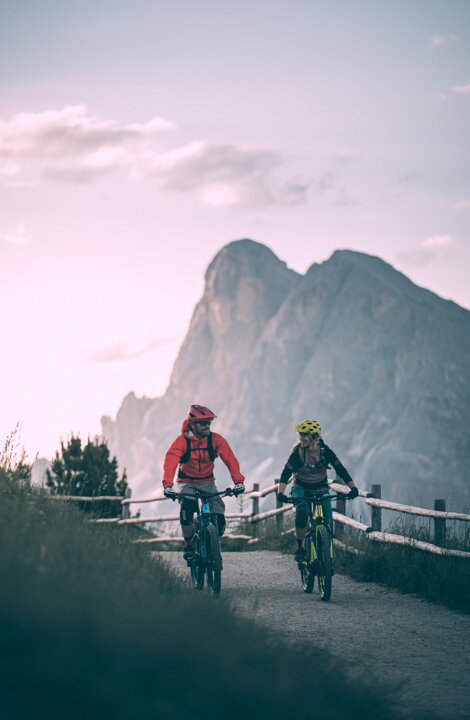 Due mountain biker pedalano su un sentiero di cresta con le montagne sullo sfondo al crepuscolo.