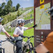 Mountain bikers check a trail map beside a route board on a sunny alpine path.