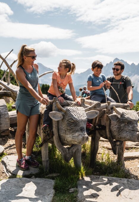 Familie mit Kindern auf einer Bergterrasse in Brixen Südtirol, auf Holzkuh-Skulpturen sitzend.