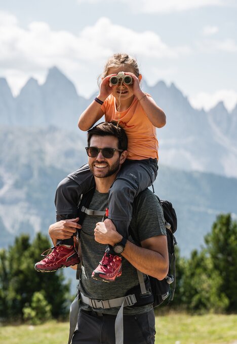 Vater trägt ein Kind auf den Schultern, im Hintergrund Berge und Himmel.