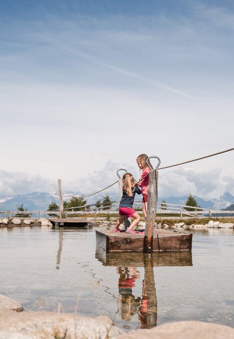 Due bambini si tengono in equilibrio su una corda sopra un laghetto poco profondo con le montagne sullo sfondo.