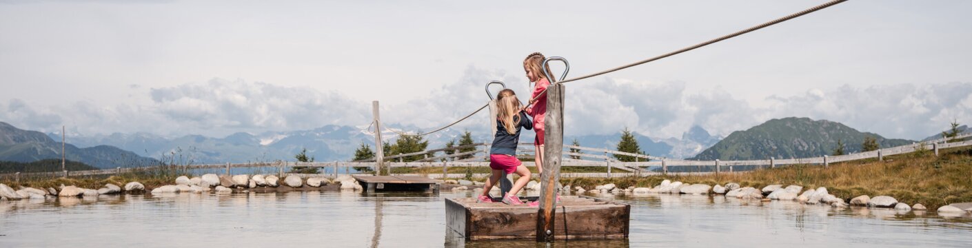 Zwei Kinder balancieren an einem Seil über einen flachen Teich mit Bergen im Hintergrund.
