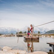 Due bambini si tengono in equilibrio su una corda sopra un laghetto poco profondo con le montagne sullo sfondo.