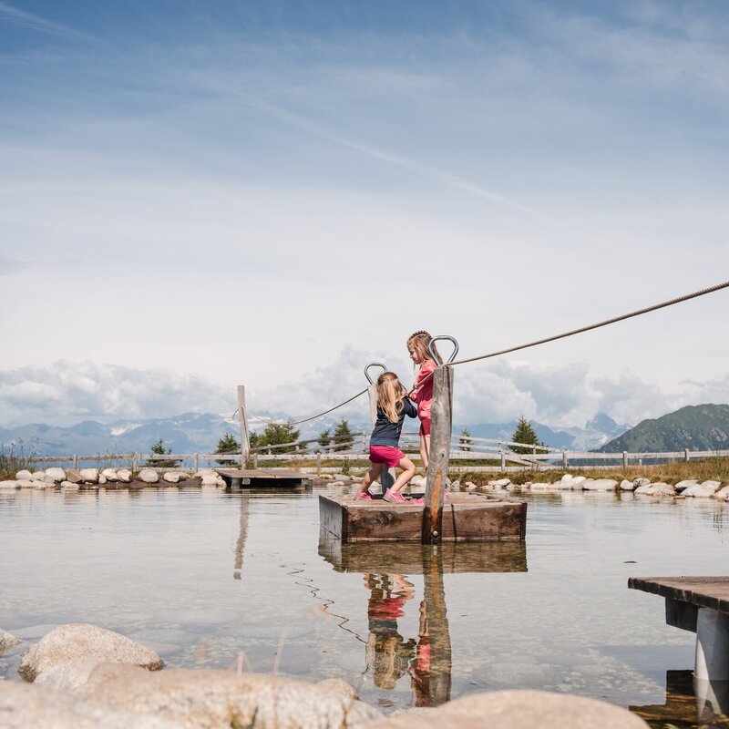 Zwei Kinder balancieren an einem Seil über einen flachen Teich mit Bergen im Hintergrund.