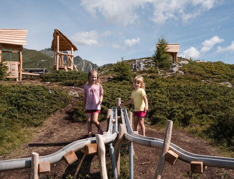 Zwei Kinder stehen auf einem Holzspielplatz an einem sonnigen Hang in den Bergen.