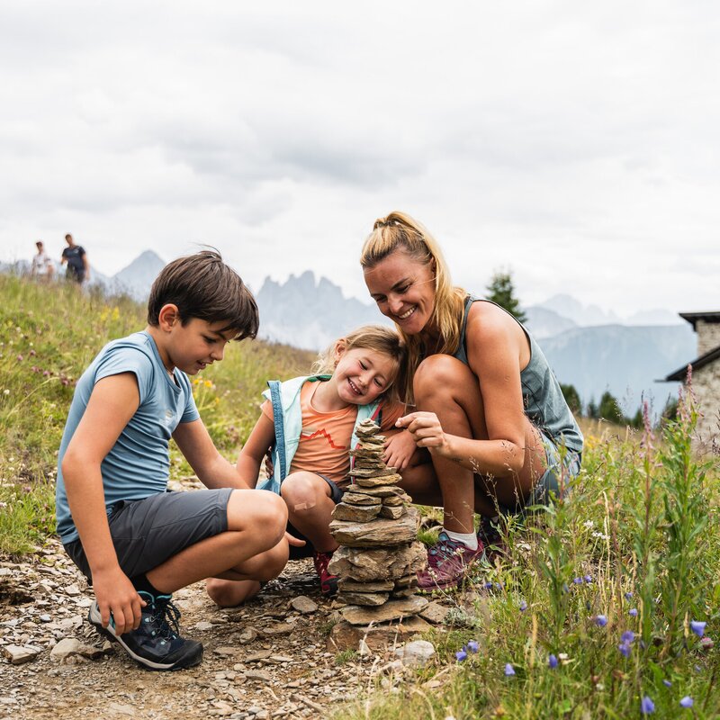 Frau und zwei Kinder bauen einen Steinturm auf einem Wanderweg in einer alpinen Wiese.
