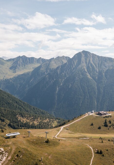 Panorama montano soleggiato con prati alpini, sentieri e cime rocciose.