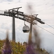 Una cabinovia sopra un prato di montagna, con cielo nuvoloso e cime alpine sullo sfondo.