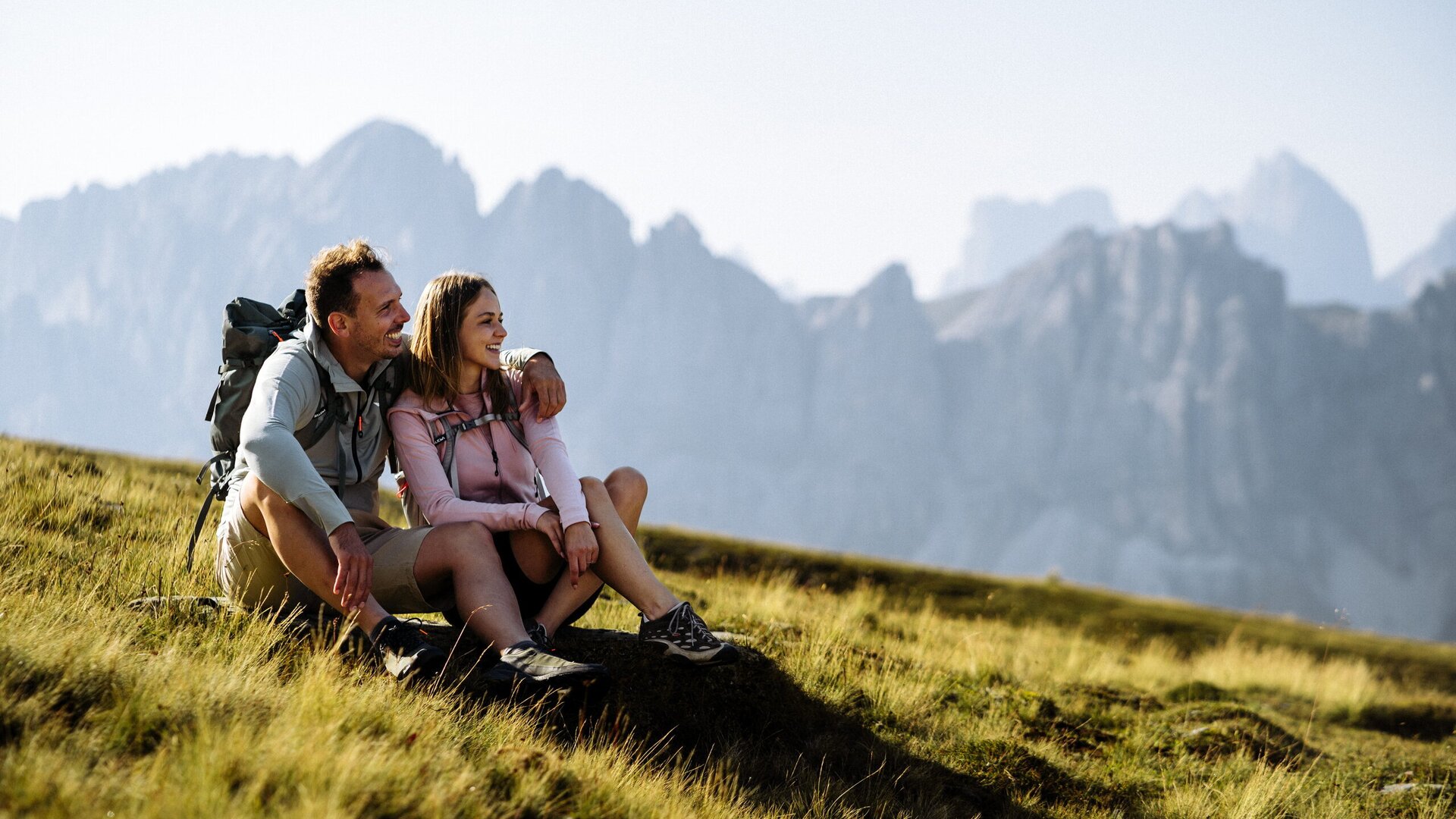 A couple rests on a grassy alpine slope with the peaks of Brixen Südtirol in the background.