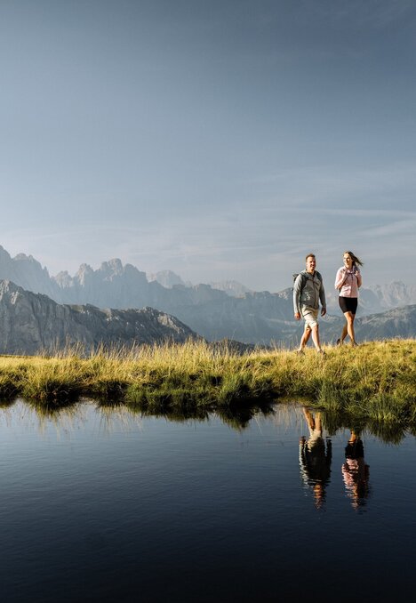 Due escursionisti camminano su un crinale erboso accanto a una pozza di montagna riflettente.