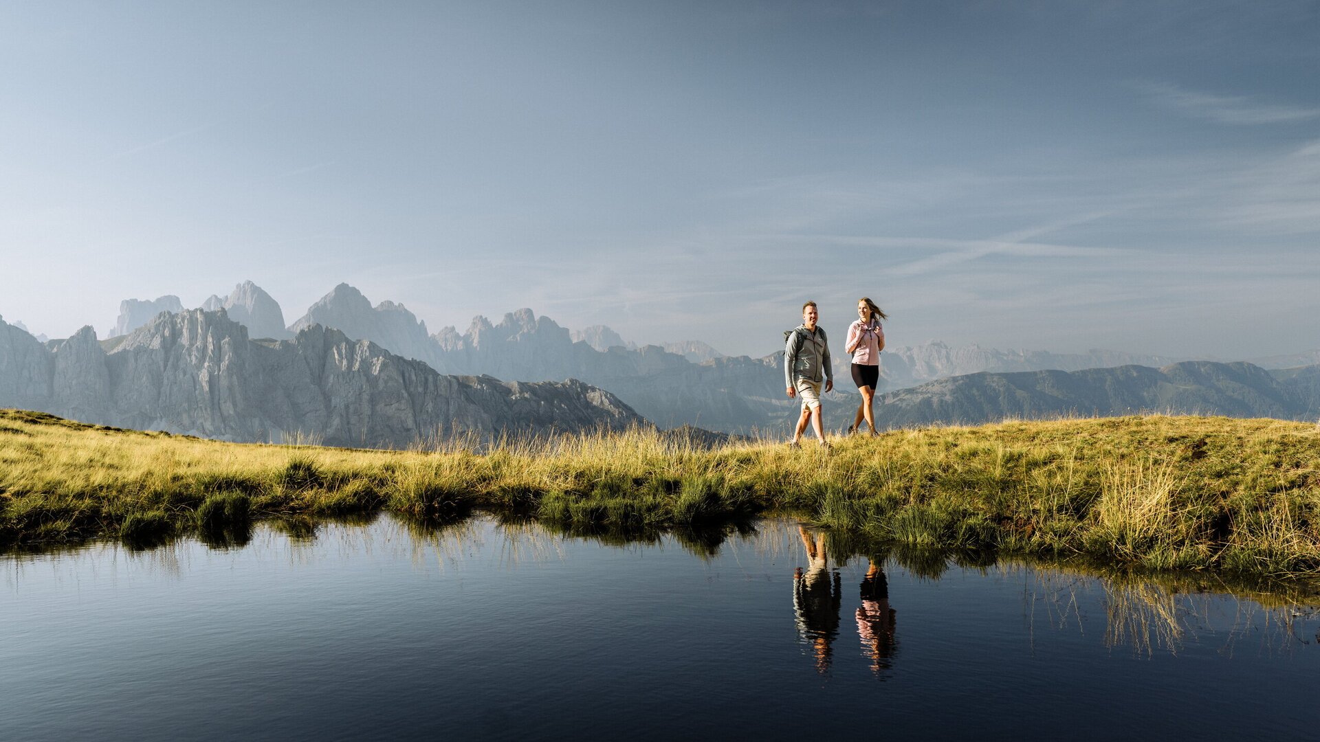 Two hikers walking along a grassy ridge beside a reflective mountain pool.