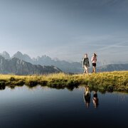 Due escursionisti camminano su un crinale erboso accanto a una pozza di montagna riflettente.