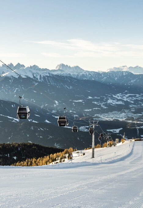 Skipiste mit Seilbahn über einem winterlichen Tal und verschneiten Berggipfeln.