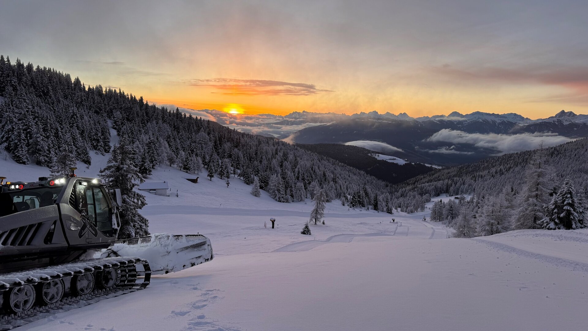 Snowcat on a snowy slope at sunrise with forested hills and distant alpine peaks.