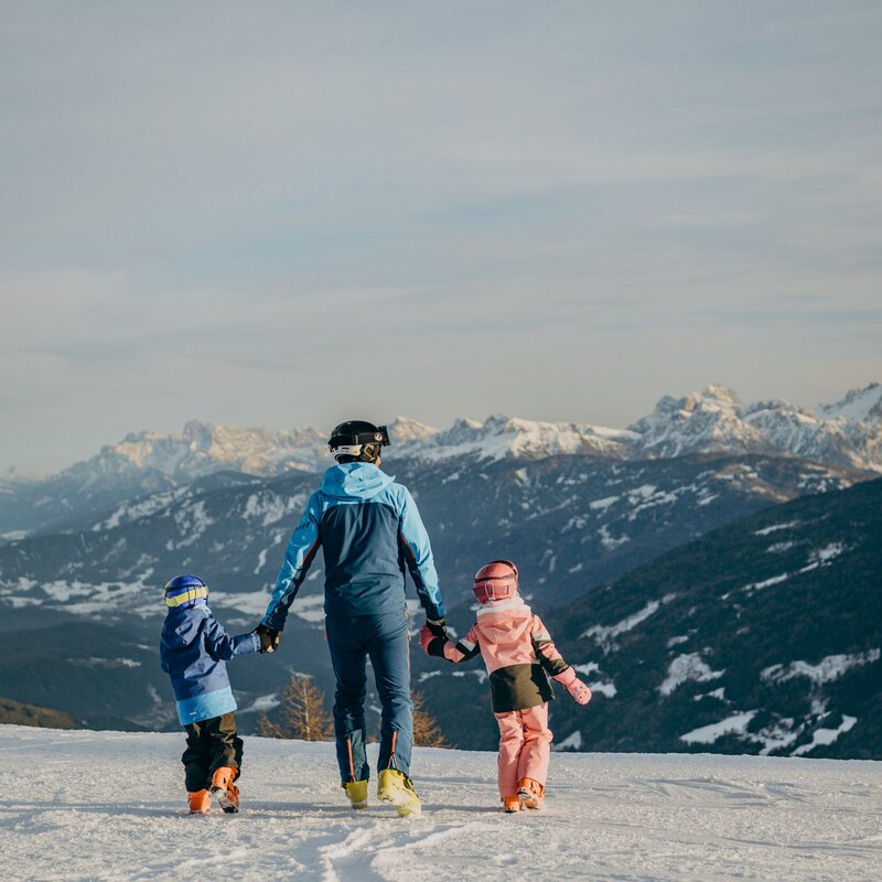 Three people walk across a snowy mountain slope with alpine peaks in the distance.