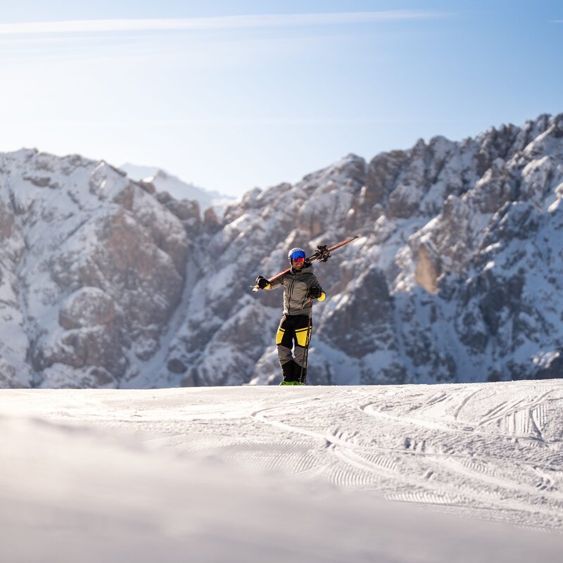 Sciatore in piedi su una pista innevata con cime alpine rocciose e cielo azzurro sullo sfondo.