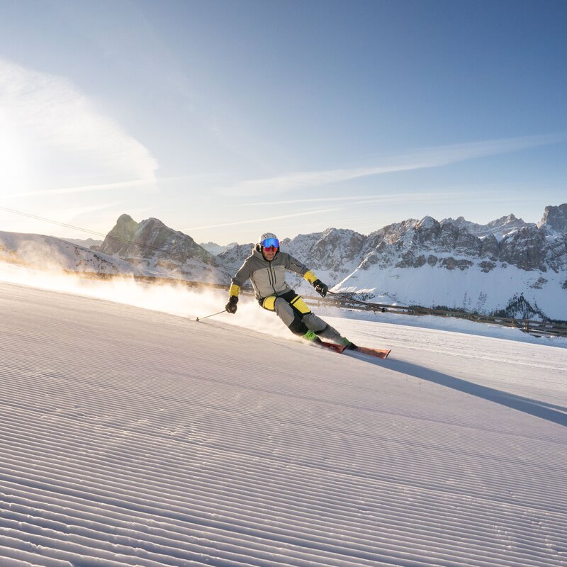 Sciatore in discesa su una pista innevata con cime montuose e sole invernale sullo sfondo.