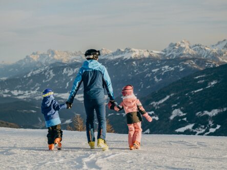 Drei Personen gehen über eine verschneite Bergflanke mit Alpen im Hintergrund.