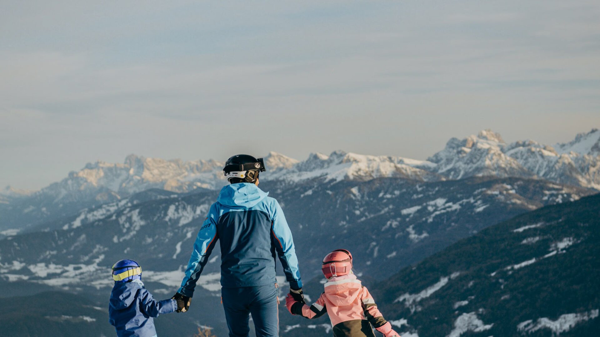 Three people walk across a snowy mountain slope with alpine peaks in the background.