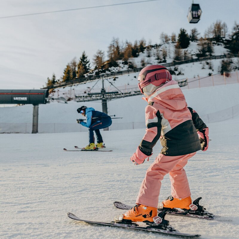 Bambino sugli sci su una pista innevata, con una funivia sullo sfondo.
