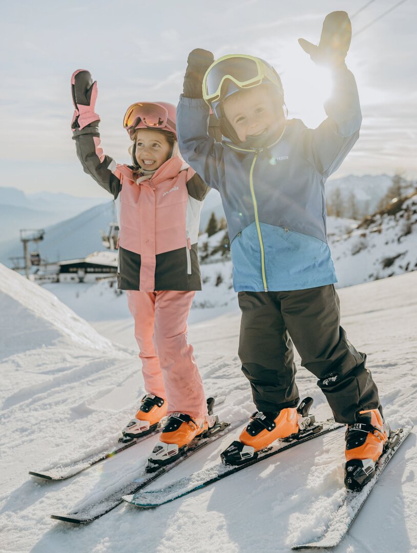 Zwei Kinder fahren gemeinsam Ski auf einer sonnigen Schneepiste in Brixen Südtirol.