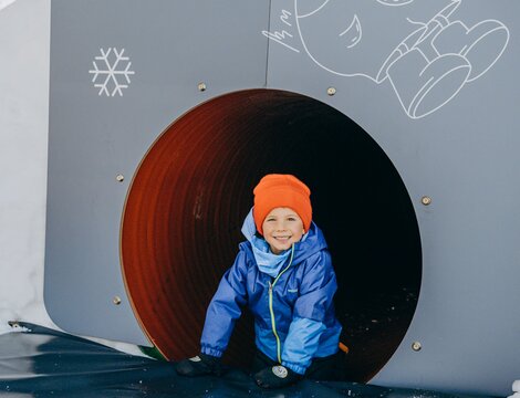 Bambino con tuta da neve blu e cappello arancione che esce da un tunnel nel GIMMY Snowpark.