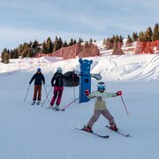 Bambini sugli sci su una pista innevata con abeti e reti rosse sotto un cielo azzurro.
