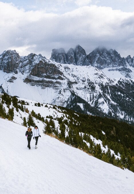 Zwei Wanderer gehen über einen verschneiten Hang vor schroffen Bergen im Wolkenhimmel.