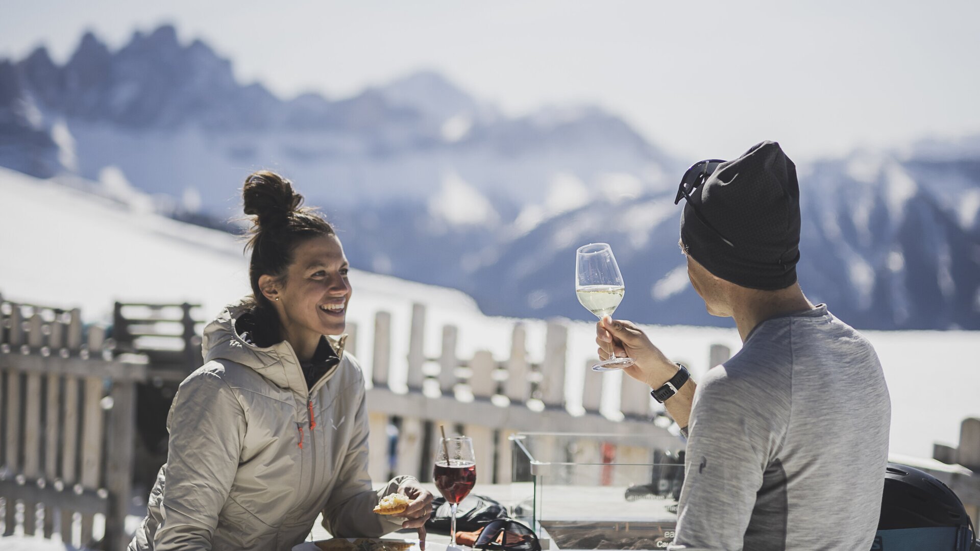 Zwei Personen essen auf einer verschneiten Terrasse mit Alpenpanorama im Hintergrund.