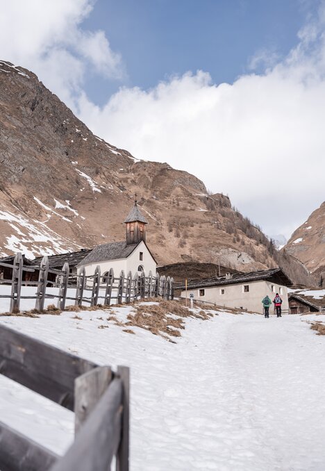Verschneiter Bergweg an einer kleinen Kirche und Almhäusern vorbei, zwei Menschen gehen in Brixen Südtirol.
