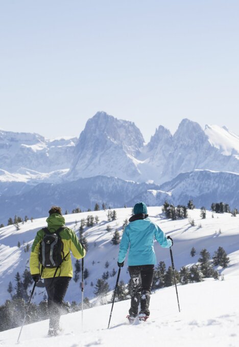 Zwei Wanderer mit Stöcken gehen über einen verschneiten Grat in Brixen Südtirol, dahinter ragen Berggipfel auf.
