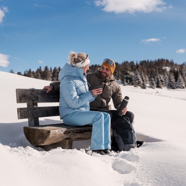 Ein Paar sitzt auf einer Bank in einer verschneiten Winterlandschaft und genießt die Sonne.