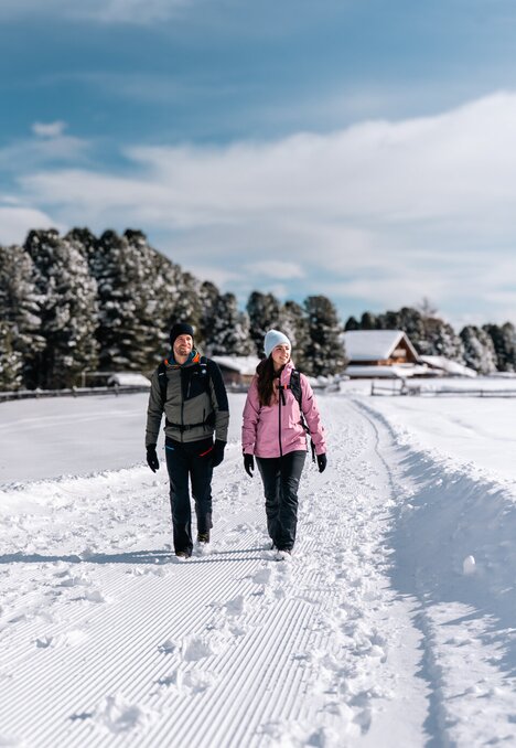 Due persone camminano su un sentiero innevato battuto a Brixen Südtirol, con alberi e una baita sullo sfondo.
