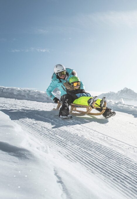 Ein Fahrer fährt mit dem Schlitten über einen verschneiten Alpenshang unter blauem Himmel.
