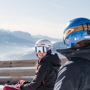 Zwei Skifahrer mit Helm sitzen auf einer Bank vor verschneiten Bergen.
