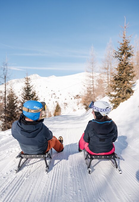 Zwei Personen fahren auf Schlitten eine verschneite Bahn unter blauem Himmel hinab.
