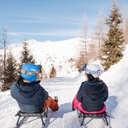 Due persone scendono su slitte lungo una pista innevata sotto il cielo blu di montagna.