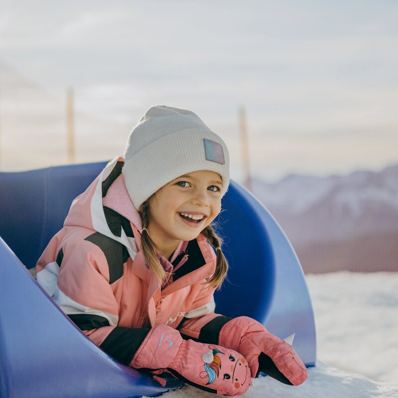 Bambina sorridente con tuta da neve rosa sdraiata sulla neve su una slitta blu