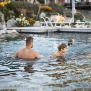 Father and child swimming in an outdoor pool with loungers and garden in the background.