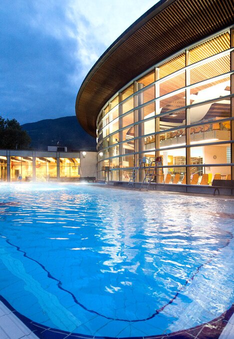 Piscina all’aperto al tramonto accanto a un moderno centro benessere con montagne sullo sfondo a Brixen Südtirol
