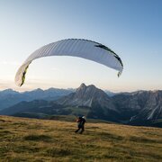 Paraglider flying above an alpine meadow with mountain peaks in the background.